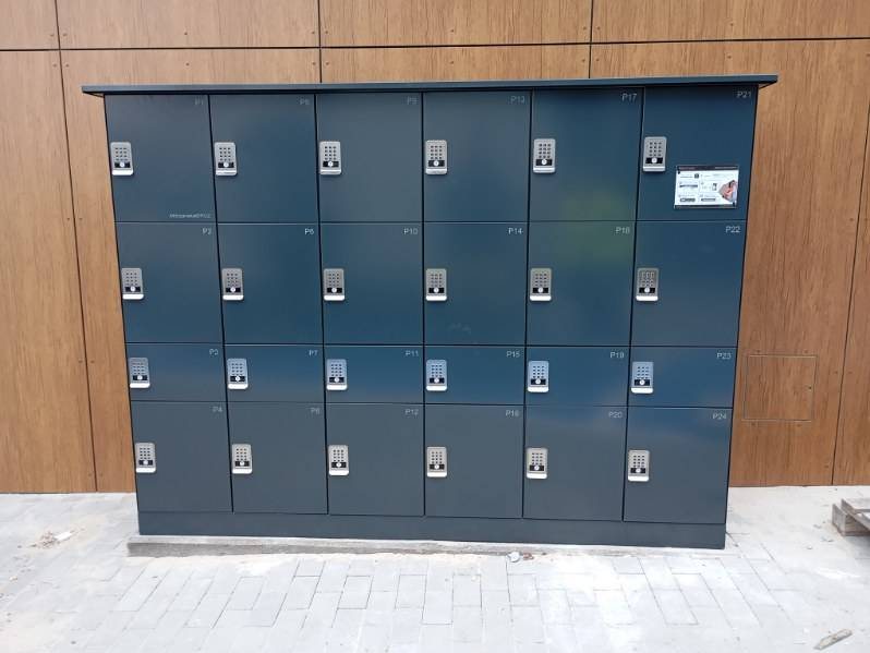 Wolnostojący grafitowy automat paczkowy Pakomatic ustawiony przy drewnianej elewacji budynku